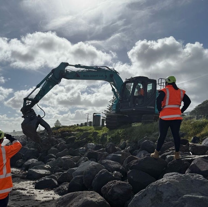 A woman in a hi-vis vest and helmet watches over a digger working on a river bank